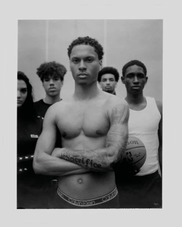 Black and white portrait of shirtless athlete posing in front of team with basketball, showcasing toned physique and ‘Sacrifice’ tattoo.
