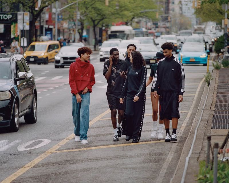 A group of young adults crossing a city street in daytime, dressed in casual athletic wear, with cars and buildings in the background.