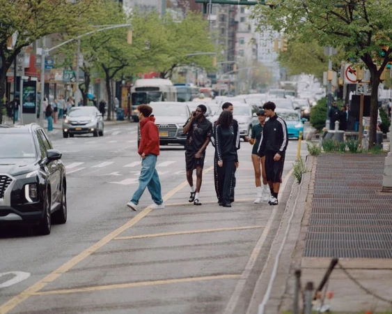 Group of casually dressed young adults cross a street in an urban setting, with cars and buildings visible in the background.