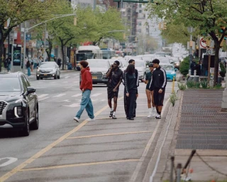 A group of models wearing athletic streetwear and tracksuits walks across a busy city street amidst traffic.