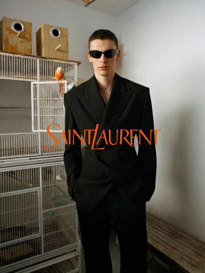 Model in a black double-breasted tailored suit and dark sunglasses standing in front of stacked birdcages.