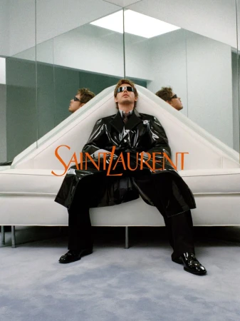 Austin Butler wearing a black patent leather trench coat and dark sunglasses seated on a white sofa for Saint Laurent.