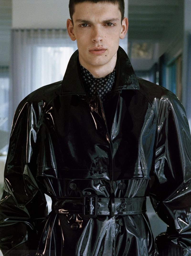 Close-up portrait of a male model wearing a glossy black trench coat and a patterned neck scarf for Saint Laurent campaign.