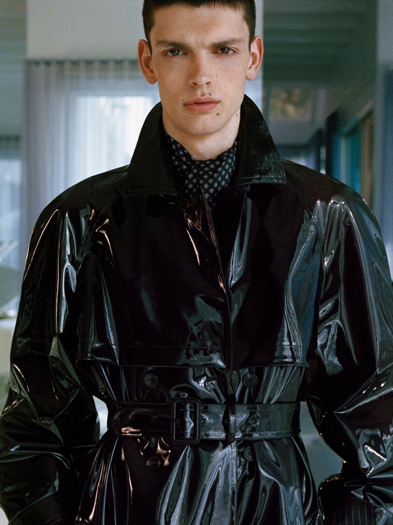 Male model wearing a high-gloss black vinyl belted trench coat with a patterned neck scarf for Saint Laurent.