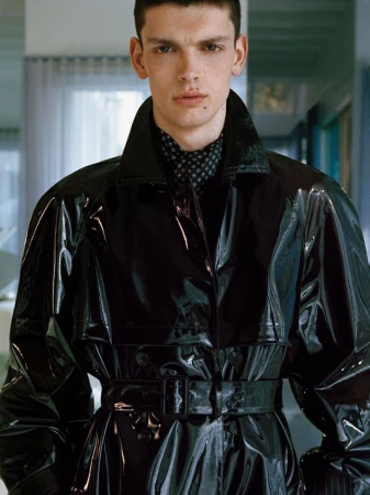 Male model wearing a high-gloss black vinyl belted trench coat with a patterned neck scarf for Saint Laurent.