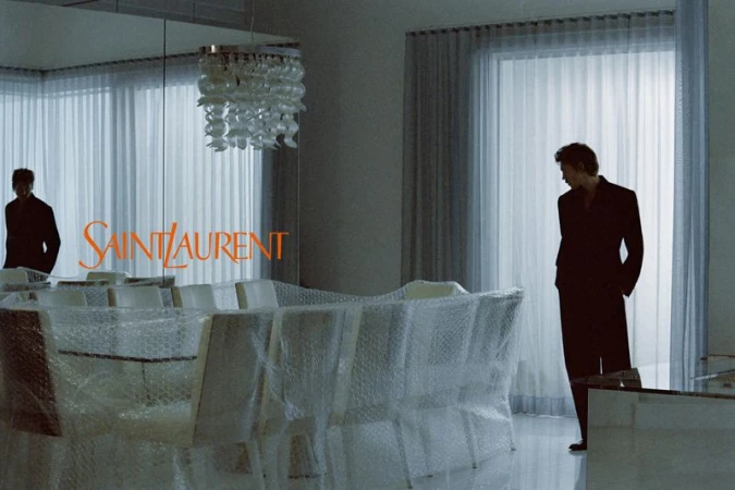 Austin Butler in a black suit standing in a room with bubble-wrapped chairs and sheer curtains for Saint Laurent.