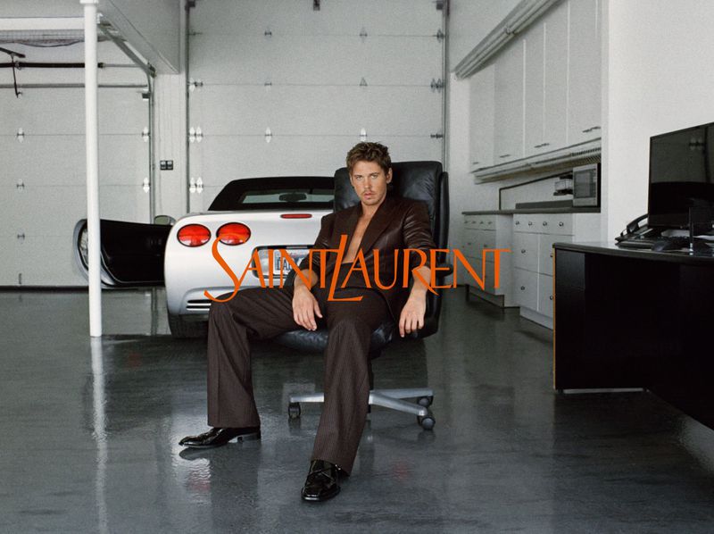 Austin Butler wearing a dark brown suit seated in an office chair in a modern garage with a silver sports car.