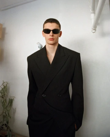 A male model in a sharp black structured blazer and dark sunglasses with a small bird on his shoulder against a plain white wall.