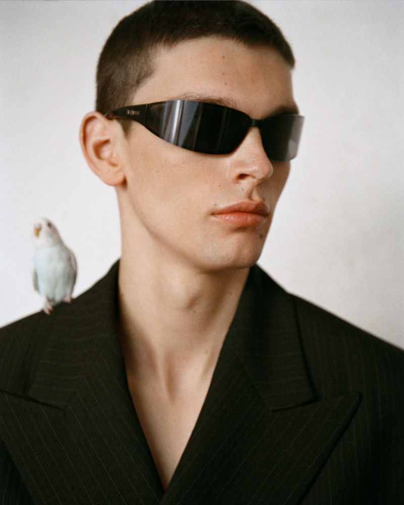 A male model wearing black sunglasses and a dark pinstripe blazer with a light blue budgerigar on his shoulder against a white wall.