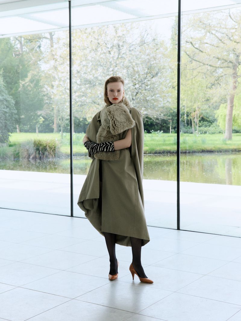Model wearing an olive green coat, fur scarf, and striped gloves standing inside a modern building with floor-to-ceiling windows.