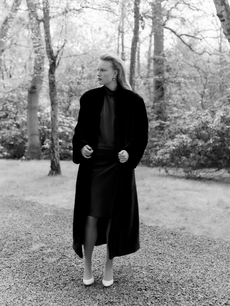 Astrid Østergaard posing in a long dark coat and white pumps on a gravel path surrounded by woodland trees.