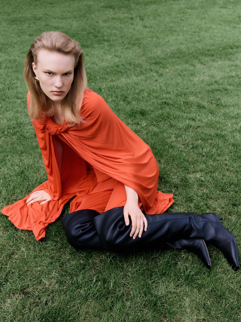 Model Astrid Østergaard wearing a draped orange dress and thigh-high black boots seated on green grass.