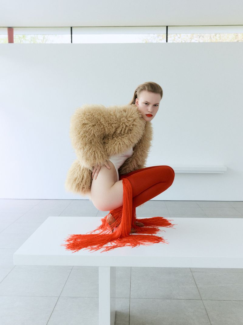 Model Astrid Østergaard crouching on a white plinth wearing a tan fuzzy jacket and red fringe tights in a bright minimalist interior.