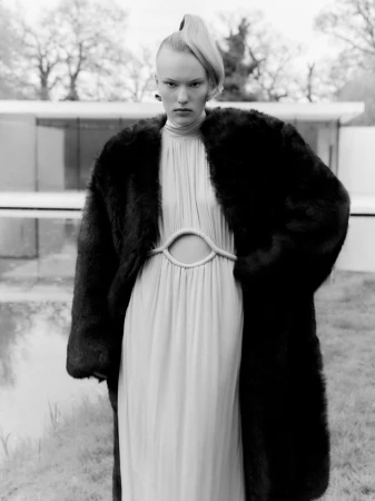 Model wearing a dark fur coat over a light pleated dress outdoors in a black and white editorial photograph