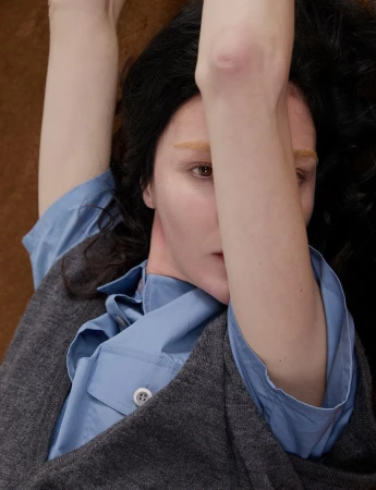 Model Mariacarla Boscono lying down in a grey knit vest and light blue shirt with her arm raised in front of her face.