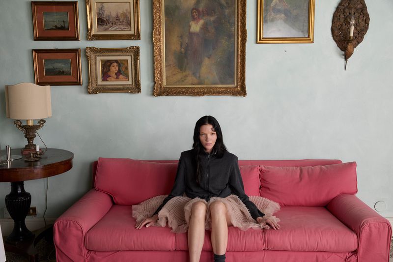 Mariacarla Boscono in a black jacket and tulle skirt seated on a pink couch beneath framed art.