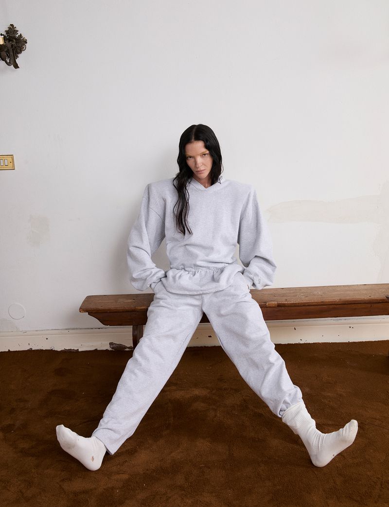 Mariacarla Boscono wearing an oversized grey sweatsuit with structured shoulders while seated on a rustic wooden bench.
