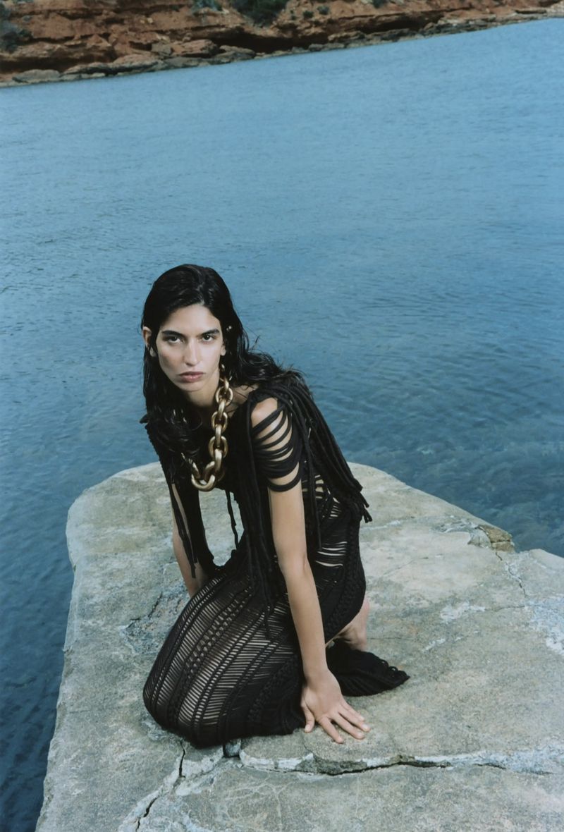 A woman with dark hair and a black dress poses on a rock formation against a blue sea backdrop with a rocky shoreline in the distance.