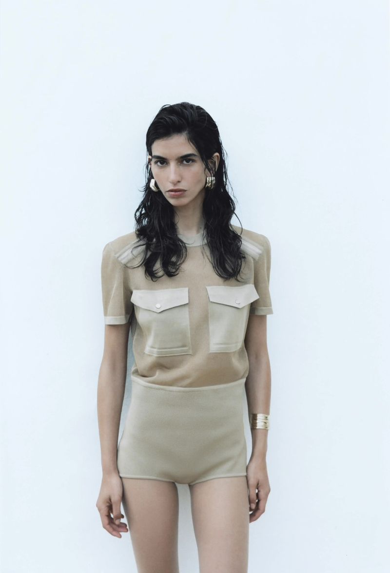 Woman in beige two-piece knit outfit with short-sleeved shirt with patch pockets and shorts, against a white background. Fashion shot.