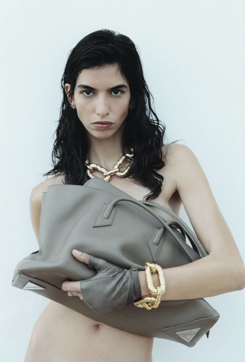 Brunette woman poses in studio wearing a gold necklace, bracelet, fingerless gloves, and holding a gray leather tote bag.