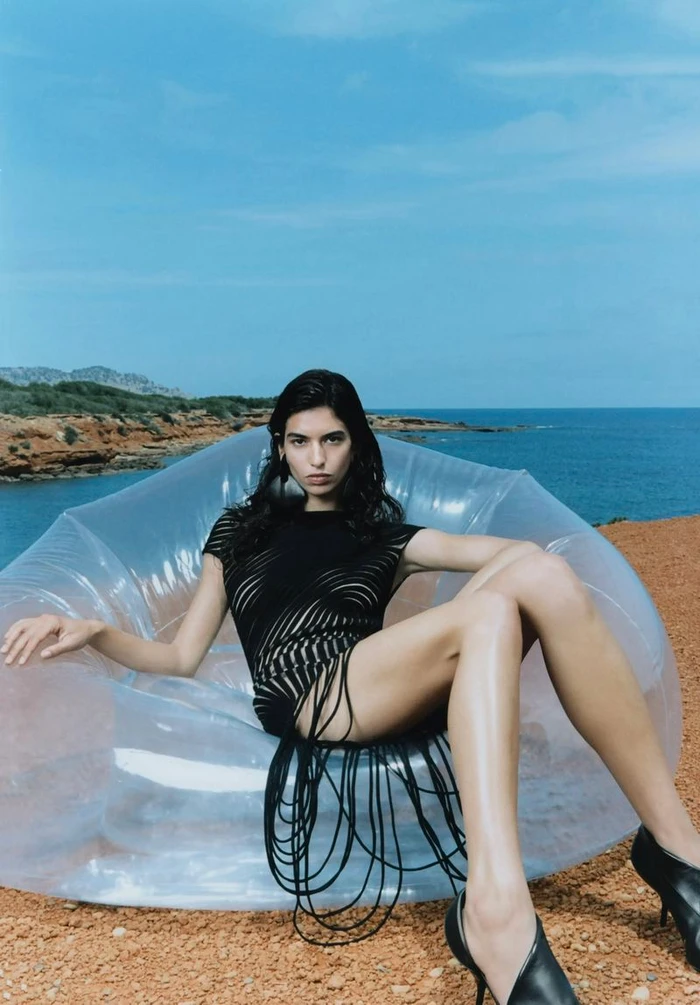 Model wearing a black cut-out dress seated in a transparent inflatable chair overlooking the sea for L'Officiel Ibiza.