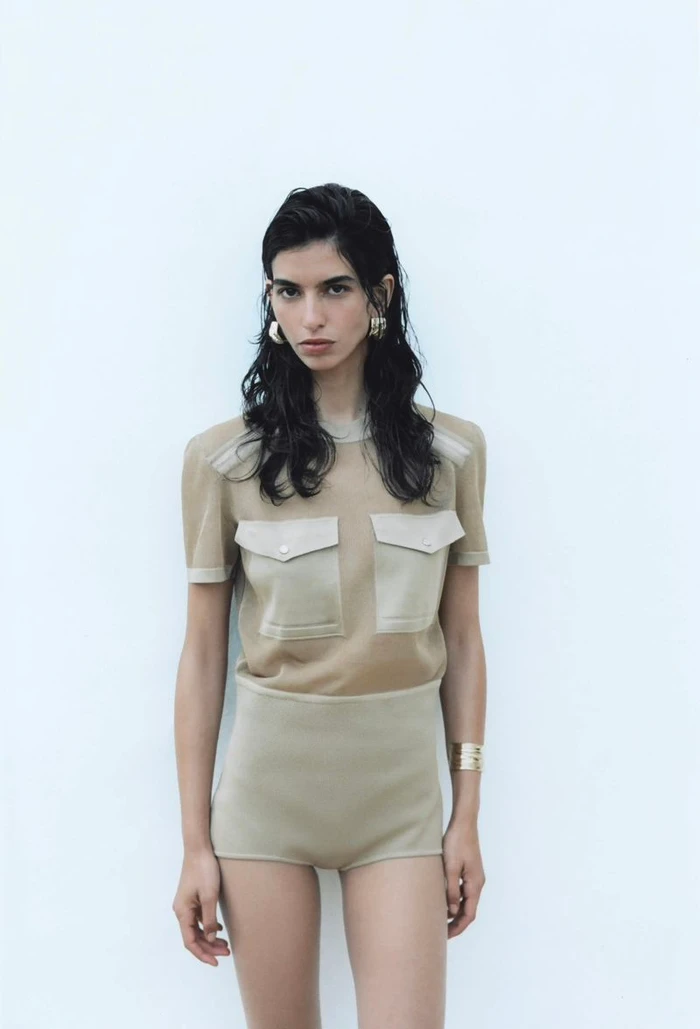 Model with wet dark hair wearing a beige short-sleeve knit top and matching shorts against a plain white background.