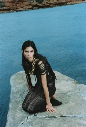 Model Gal Dahan kneeling on a rock ledge over coastal water in a black open-knit dress and chunky gold chain necklace.
