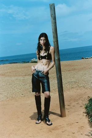 Model Gal Dahan posing by a wooden pole on a sandy beach wearing a black crop top, leather shorts, and black boots.