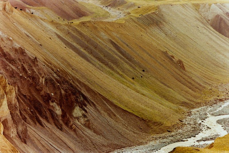 Steep layered mountain slopes in earthy ochre and russet tones with a winding stony riverbed at the valley base.