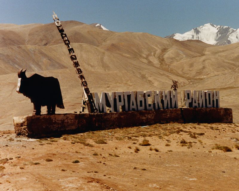 Cyrillic roadside monument with a yak sculpture against arid desert terrain and snowcapped mountains in the Murghab District.