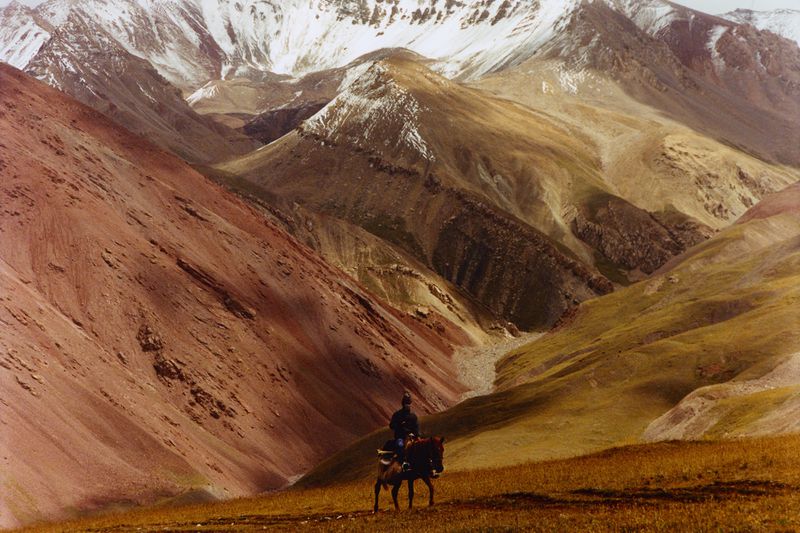 Horseback rider traversing a golden grassy slope with reddish hills and snow-dusted mountains in the background.