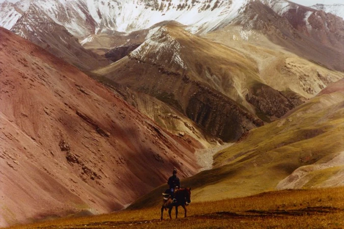 Horseback rider traversing a golden grassy slope with reddish hills and snow-dusted mountains in the background.