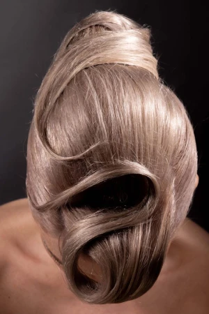 High-angle view of an elaborate sculptural blonde hair updo with glossy swirling locks against a dark studio backdrop.