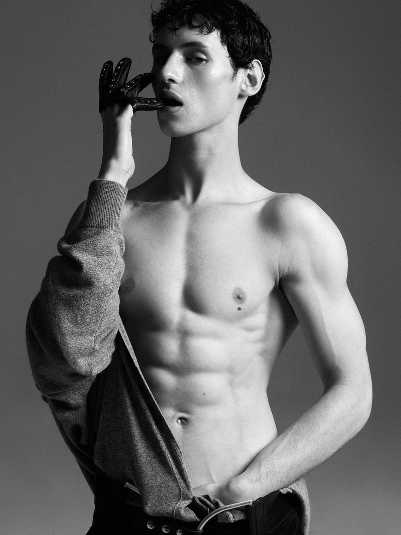Shirtless male model wearing a studded black leather glove and a draped grey sweatshirt against a grey studio backdrop.