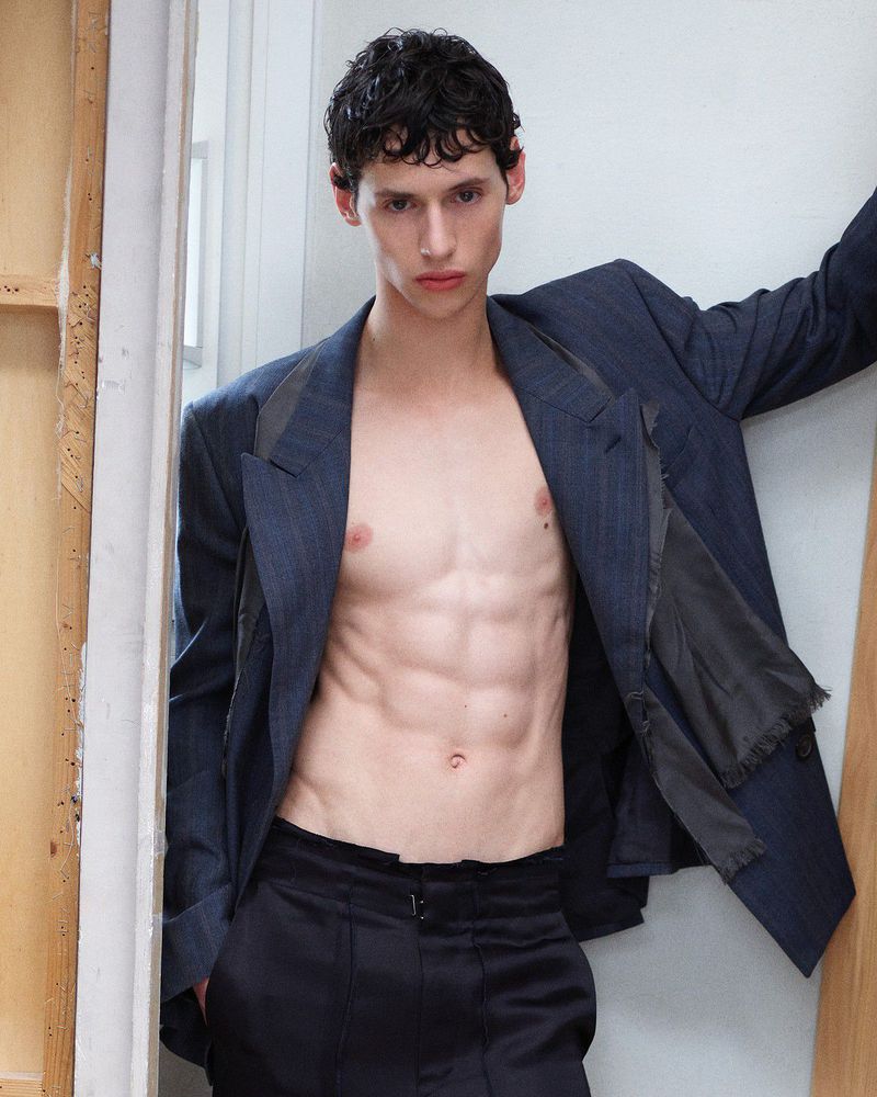 Shirtless male model wearing an open oversized navy blazer and black trousers leaning against a studio wall.