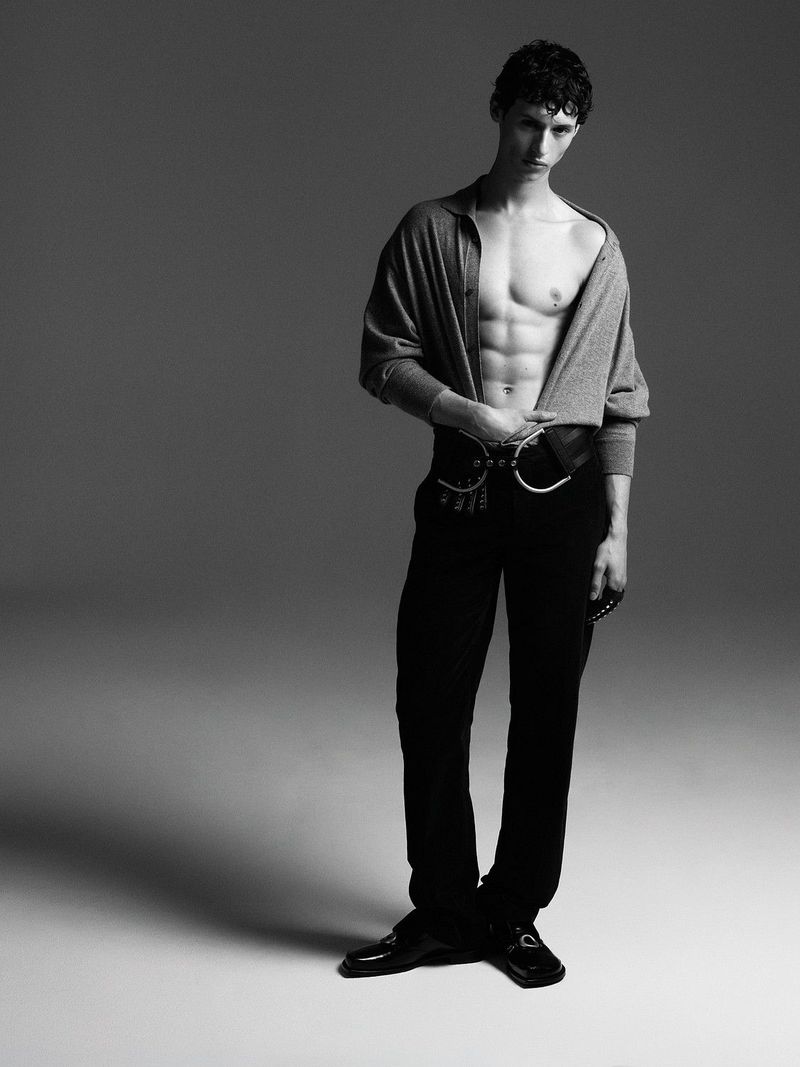Model Paolo Borle posing shirtless in an open grey knit top and black trousers with an ornate belt against a grey studio backdrop.