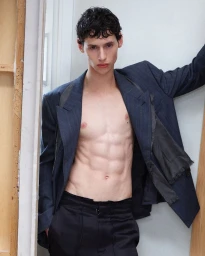 Shirtless male model wearing an open oversized navy blazer and black trousers leaning against a studio wall.