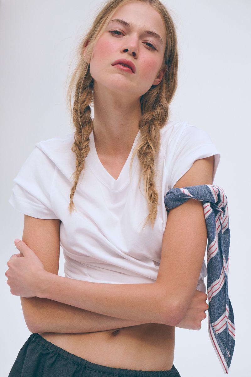 Model Saphira Krumhaar posing in a white v-neck t-shirt with braided pigtails against a white studio background.