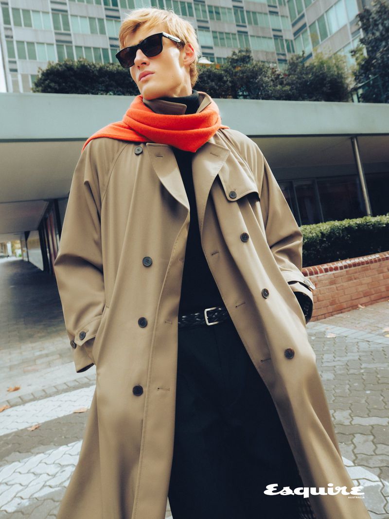Man wearing a camel trench coat, black sunglasses, and a bright orange scarf outdoors with modern buildings in the background.