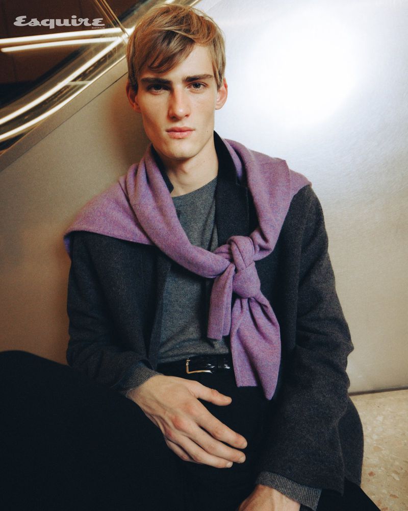Young man seated wearing a charcoal jacket with a lilac sweater knotted around his shoulders for Esquire Australia.