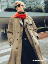 Man wearing a camel trench coat, black sunglasses, and a bright orange scarf outdoors with modern buildings in the background.