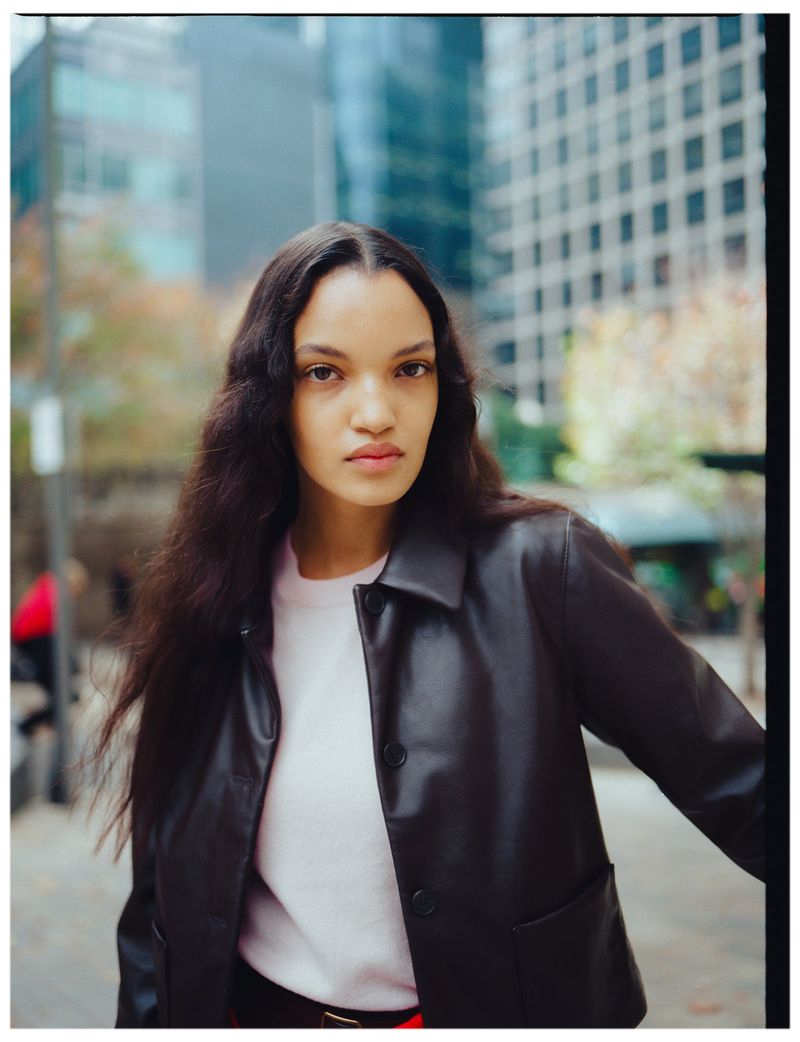 Model in a brown leather jacket and pale pink sweater posing outdoors on a city street in front of office buildings.