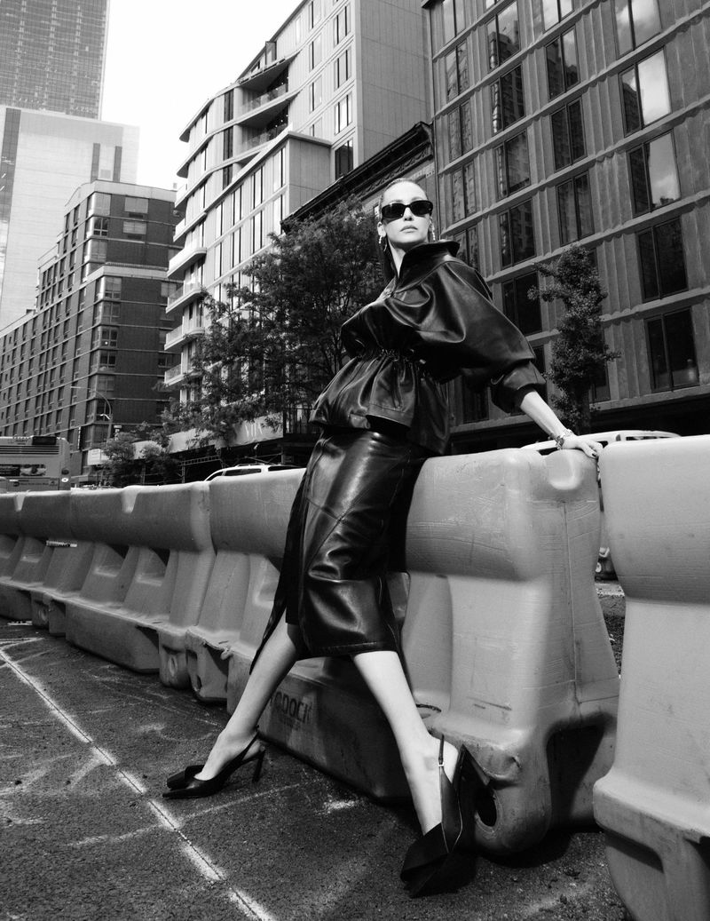 Nina Dobrev leaning against a plastic road barrier in an urban street setting wearing dark sunglasses and a leather skirt and jacket.