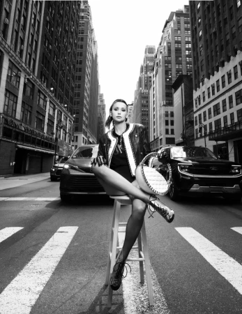 Nina Dobrev sits on a tall white wooden stool in a crosswalk on a city street flanked by skyscrapers in black and white.