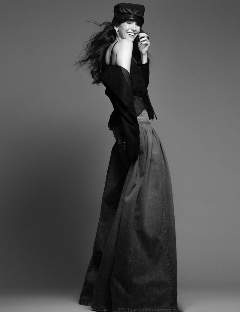 Nina Dobrev smiling in a black cap, off-shoulder black top, and voluminous wide-leg denim maxi skirt against a neutral studio backdrop.