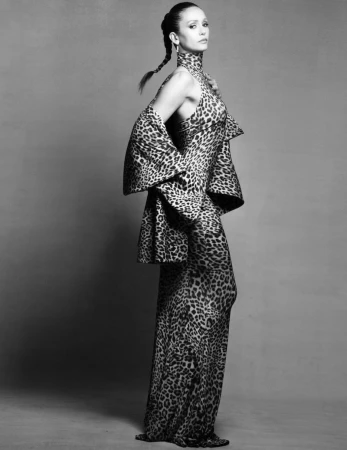 Nina Dobrev posing in a full-length leopard-print high-neck dress and cape against a neutral studio backdrop.