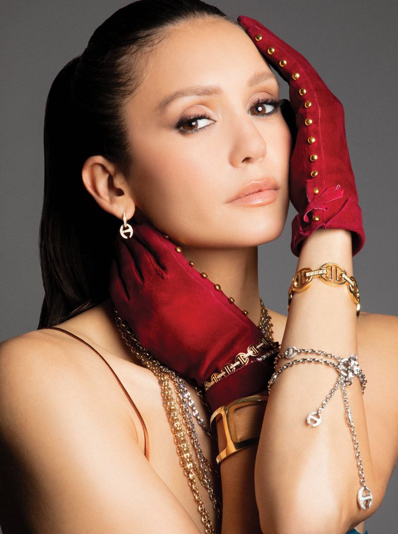 Nina Dobrev wearing red studded gloves and layered gold and silver bracelets posing against a grey studio backdrop.