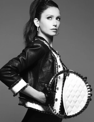 Nina Dobrev wearing a black cropped leather jacket and holding a circular white quilted handbag against a grey studio backdrop.