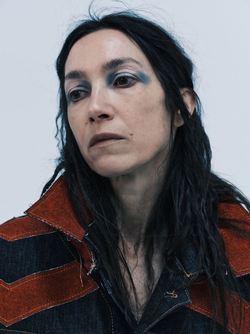Close-up portrait of a woman with smoky blue eyeshadow wearing a dark denim jacket with rust chevron details against a plain background.