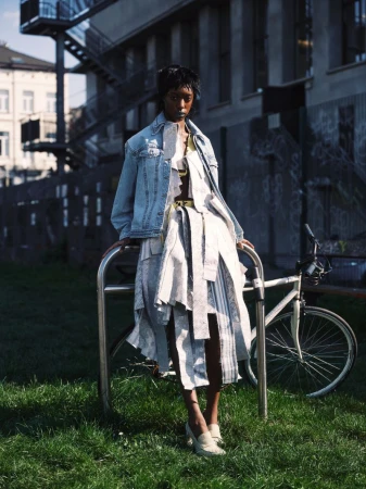 Model wearing a light-wash denim jacket and a deconstructed layered tie dress posing next to a bicycle on green grass.